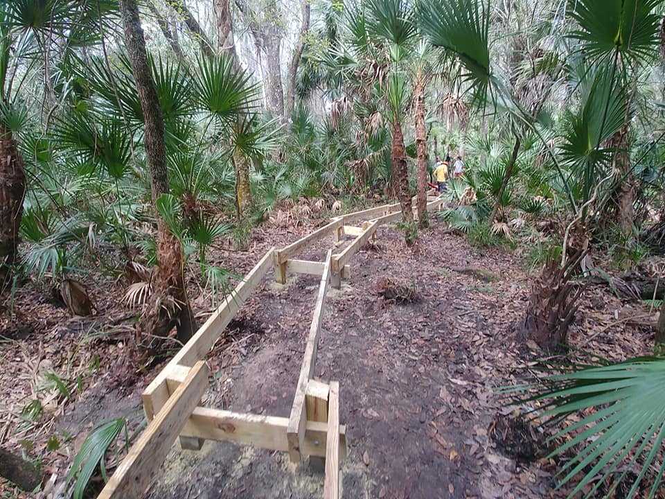 Boardwalk framing — pressure-treated lumber laid out through the wetland with volunteers visible in the background