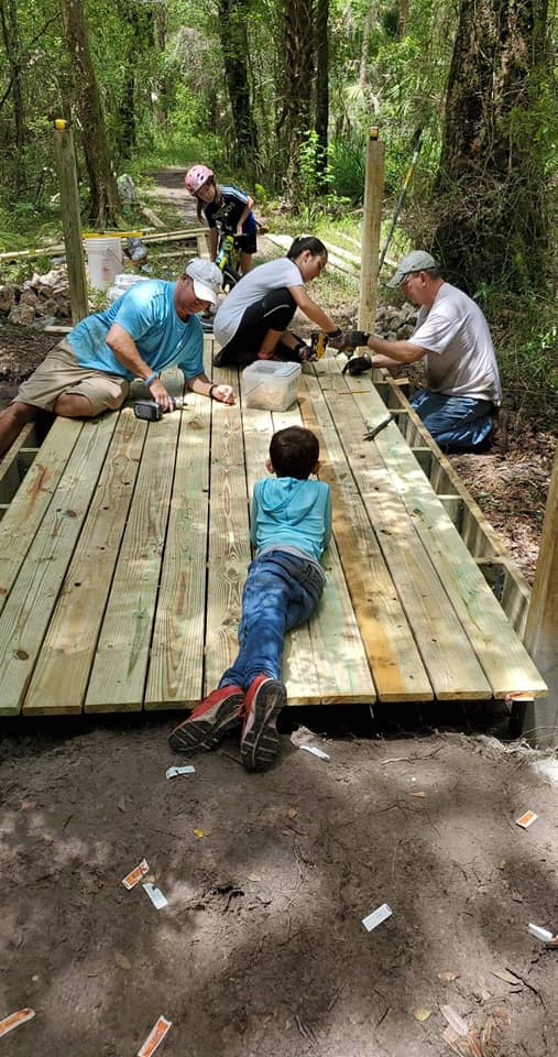 Volunteer crew building Bridge #3 — including a young child lying flat on the deck helping screw down boards