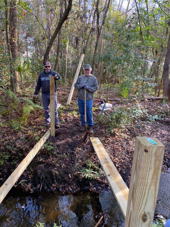 Two volunteers posing with the first beam spanning the creek — early framing of Cordgrass Bridge