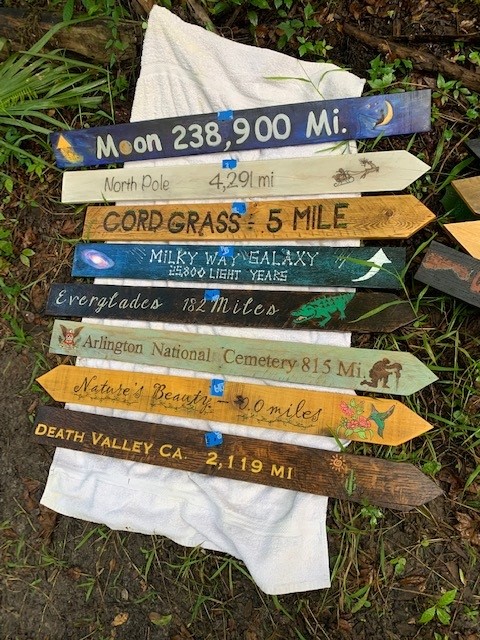 All 15 hand-painted directional signs laid out on the ground — Moon 238,900 Mi., North Pole, Milky Way, Everglades, Arlington National Cemetery, Death Valley, and more
